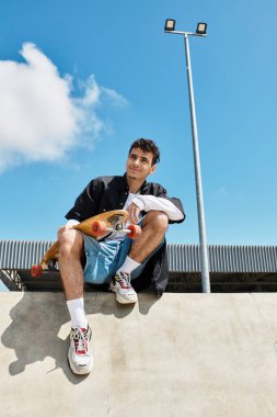 Genç bir adam, mavi gökyüzünün altında kaykayını tutarak parktaki bir çıkıntıda dinleniyor..