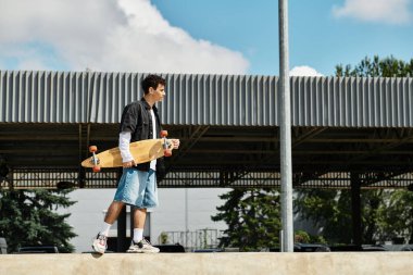 Genç bir adam elinde kaykayıyla parkta geziniyor, açık havada güneşli bir günün tadını çıkarıyor..