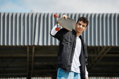 Genç bir adam parkta kaykayını tutarak güzel gökyüzüne gülümsüyor..