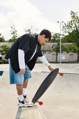 Gündelik kıyafet giymiş genç bir adam güneşli bir park ortamında kaykayına odaklanmış..