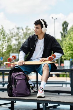 Genç bir adam parkta dinleniyor, kulaklık takıyor ve elinde kaykayla günün tadını çıkarıyor..