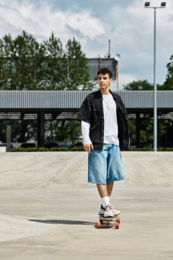 Genç bir adam kaykayının üstünde duruyor, yeteneğini canlı bir park atmosferinde gösteriyor..