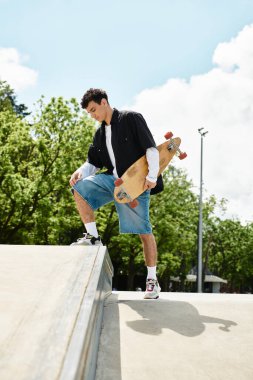 Genç bir adam parlak bir parktaki kaykay rampasında duruyor, açık gökyüzünün altında sürüşe hazırlanıyor..