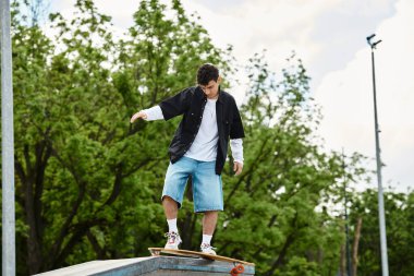 Genç adam parkta kaykayın üstünde dengede duruyor, uzun ağaçlar arasında becerilerini gösteriyor..