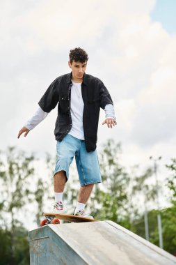 Genç bir adam açık bir günde kaykayıyla parktaki rampada ustalıkla dengede duruyor..