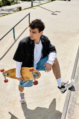 Kıvırcık saçlı genç bir adam güvenle parkta duruyor, elinde kaykayıyla..