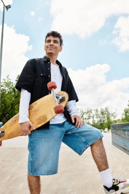 Genç bir adam parkta elinde kaykayla rahatlar ve keyifli havanın tadını çıkarırken gülümser..