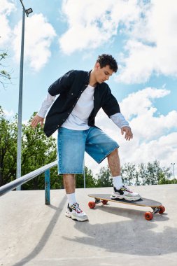 Genç bir adam parlak mavi gökyüzünün altındaki canlı bir parkta kaykay yeteneğini gösteriyor..