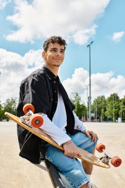 Genç bir adam parktaki bankta rahat bir şekilde oturur ve kaykayını neşeli bir ifadeyle gösterir..