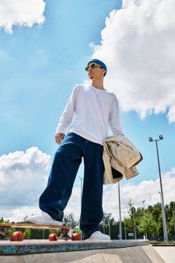 Parkta parlak bir gökyüzünün altında kaykayın üstünde duran genç bir adam gündelik tarzıyla hava atıyor..