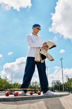 Genç bir adam kaykay parkında duruyor, güneş gözlüğü takıyor ve ceketini tutuyor, eğlenceye hazır..