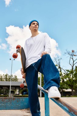 Genç bir adam güvenle parkta duruyor, kaykayını tutuyor ve güneşi emiyor..