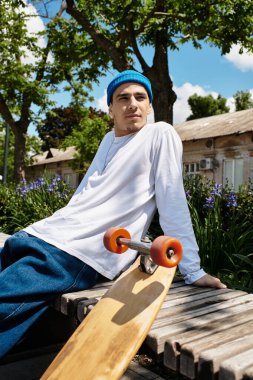 Genç bir adam güneşin altındaki bir bankta dinleniyor, yanındaki kaykayla park atmosferinin tadını çıkarıyor..