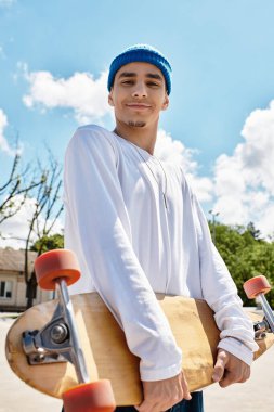 Genç bir adam açık mavi gökyüzünün altında neşeyle bir kaykay tutuyor..