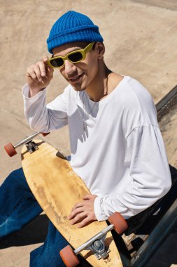 Genç bir adam parkta oturuyor, güneş gözlüğü ve beresi var, yanında kaykayıyla..
