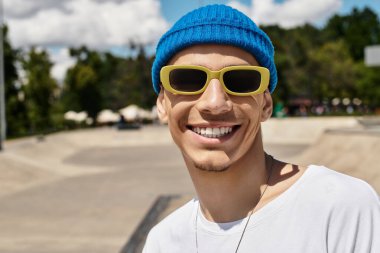 Neşeli genç bir adam, parlak bir günde, bereli ve güneş gözlüklü, güven içinde parkta poz verir..