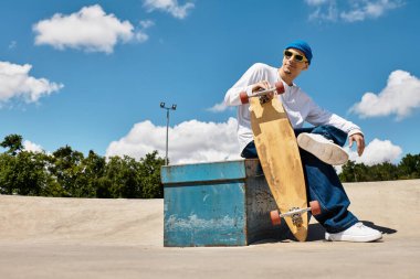 Parktaki mavi bir kulübede oturan genç bir adam parlak gökyüzünün altında kaykayını gösteriyor..