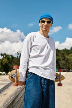 Genç adam kaykay parkında duruyor, kaykayını canlı mavi gökyüzünün altında tutuyor..