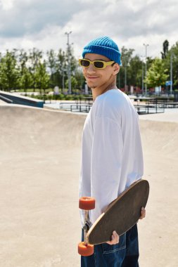 Genç bir adam parkta gülümsüyor, kaykayını tutuyor ve günlük kıyafetlerini giyiyor..