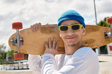 Neşeli genç bir adam parkta güneşin altında parlak bir şekilde gülümserken kaykayını tutuyor..