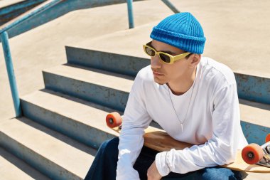 Genç bir adam, mavi bir bere ve güneş gözlüğü takarak parktaki basamaklarda rahatlamak için biraz zaman ayırır..