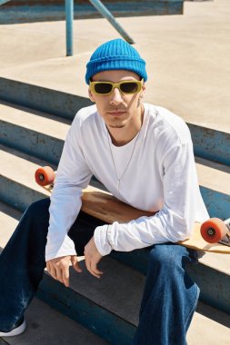 Bir adam park basamaklarında, güneş gözlüğü ve parlak bir bere takıyor, güneşin tadını çıkarıyor..