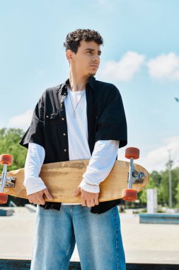 Genç bir adam güvenle parkta duruyor, kaykayını tutuyor ve uzaklığa bakıyor..