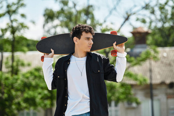 Молодой человек уверенно стоит в парке со скейтбордом, глядя вдаль.