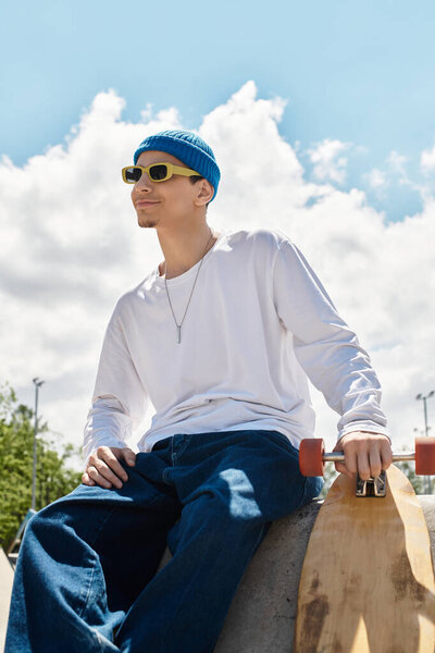 Bright sunlight shines on a young man dressed casually, sitting with a skateboard at the park.