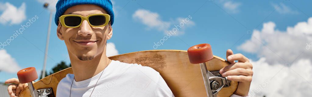 A young man smiles while holding a skateboard in the park under a clear blue sky.