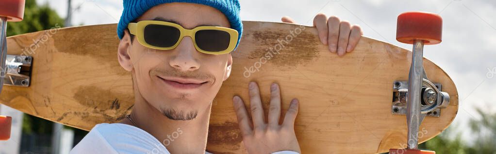 A young man smiles while holding his skateboard in the park, enjoying a bright sunny afternoon.