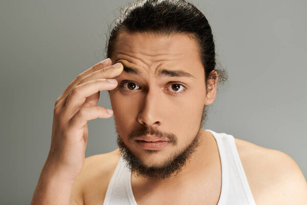 A young man with a beard looks puzzled while touching his forehead in a well lit studio.
