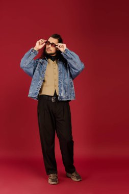 Sakallı yakışıklı genç bir adam gündelik kıyafetlerini gösterirken güneş gözlüğünü ayarlıyor..