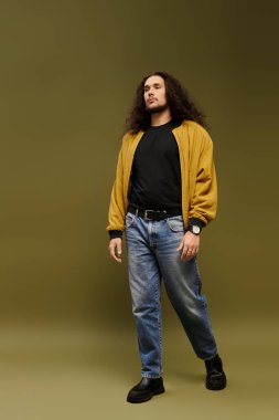 Sakallı ve kıvırcık saçlı genç bir adam güvenli bir şekilde yürüyor, ceket giyiyor ve rahat kot pantolon giyiyor..