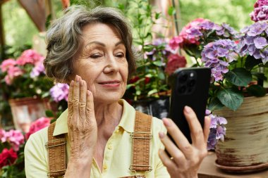 Neşeli yaşlı bir kadın renkli bahçe çiçekleriyle çevriliyken akıllı telefonuna hayran kalır..