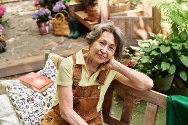 Yaşlı bir kadın çiçek ve bitkilerle dolu güzel bir bahçede bir bankta dinleniyor..