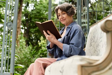 Huzurlu bir bahçede, neşeli bir yüzü olan yaşlı bir kadın rahatça otururken kitap okuyor..
