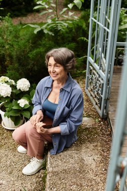 Güzel, yaşlı bir kadın çiçek ve yeşilliklerle çevrili bahçe yolunda dinleniyor..