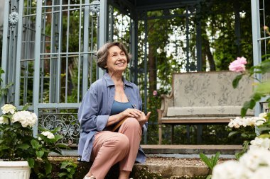 Gülümseyen, son sınıf öğrencisi bir kadın yemyeşil bir bahçede huzur içinde oturur..