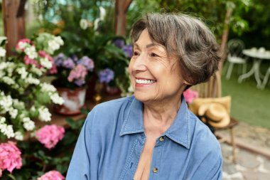 Son sınıf öğrencisi bir kadın, yemyeşil bir bahçede renkli çiçeklerle çevrili bir şekilde gülümsüyor..