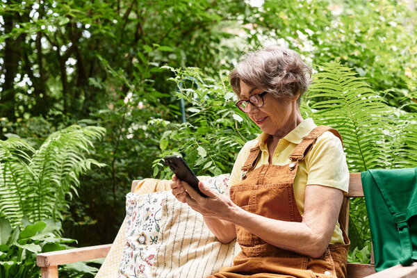 Beautiful senior woman relaxes in a lush garden, engaging with her smartphone in a tranquil moment.