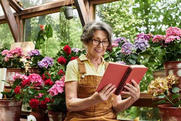 A beautiful senior woman smiles while reading a book surrounded by blooming plants and flowers.