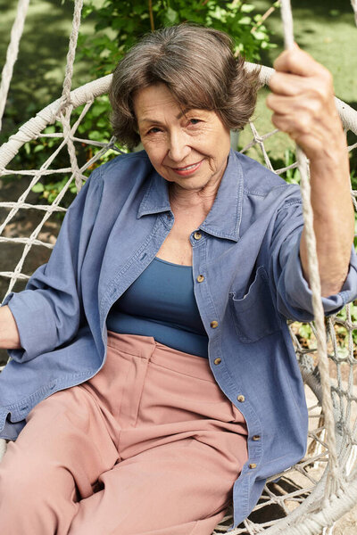 A senior woman sits comfortably on a swing, surrounded by the tranquility of a flourishing garden.