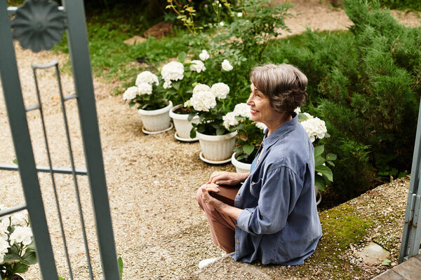 A senior woman smiles while sitting in a tranquil garden surrounded by blooming flowers.