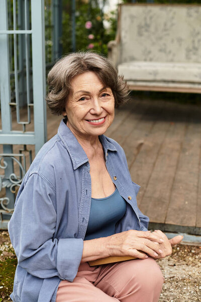 A senior woman kneels joyfully in her lush garden, surrounded by vibrant plants and tranquility.