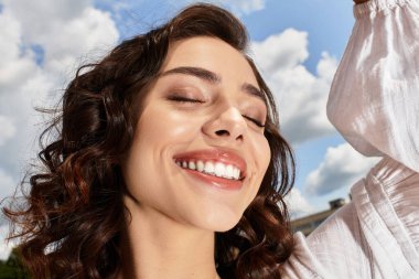 Dalgalı kahverengi saçlı güzel bir kadın güneşli bir yaz gününde neşeyle gülümsüyor..