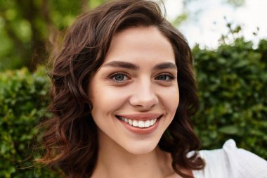 Dalgalı kahverengi saçlı güzel bir kadın güneşli bir parkta dururken neşeyle gülümsüyor..