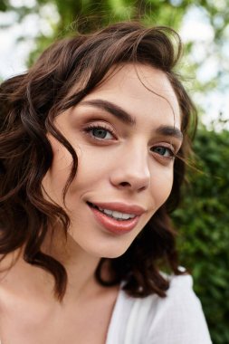 Dalgalı kahverengi saçlı güzel bir kadın parkta otururken ışıl ışıl gülümsüyor..