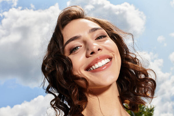 Красивая молодая женщина с волнистыми каштановыми волосами тепло улыбается на открытом воздухе в яркий летний день.