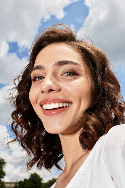 A young woman with wavy brown hair enjoys a summer day, smiling under the blue sky while outdoors.